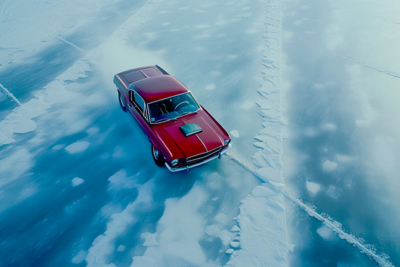 A crimson 1960s muscle car with a hood scoop drives across a stark, icy blue landscape, showcasing power and elegance in winter conditions.